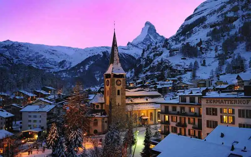  Nighttime view of the cozy village of Zermatt against a pink sky, embraced by snowy mountains, emanating a tranquil and charming winter ambiance.